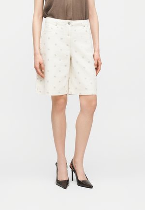 Γυναίκα που φοράει λευκά σορτς έως το γόνατο με μικρά ανθισμένα σχέδια, ένα καφέ τοπ και μαύρα μυτερά ψηλοτάκουνα παπούτσια.