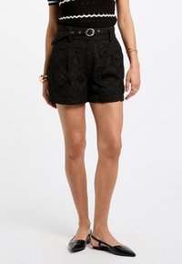 Black shorts with floral lace texture, high waist, pleats, and a matching belt featuring a silver buckle. Styled with pointed black flats.