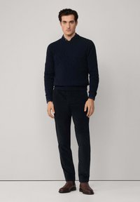 Maglione a trecce navy, pantaloni in velluto a coste blu scuro e stivali in pelle marrone. Modello in piedi diritto, mani ai fianchi, sfondo minimalista.