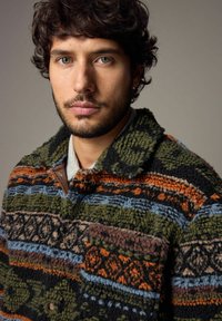 Fuzzy, multikleur pullover jack met groene, oranje, blauwe en zwarte patronen, een kraag met knopen en een gestructureerd oppervlak.