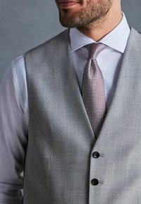 Gilet de costume gris avec une finition texturée, comportant deux boutons noirs. En dessous, une chemise blanche et une cravate à motifs rose.