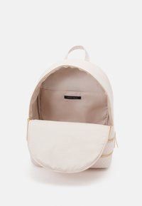 Mochila de color beige claro con cremalleras doradas, forma redondeada, base texturizada y un interior suave. Cuenta con un compartimento grande y un bolsillo pequeño.
