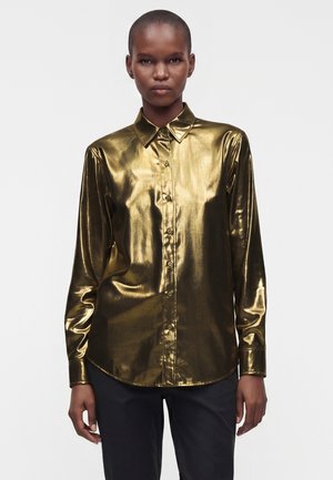 Persona che indossa una camicia dorata lucida a maniche lunghe con bottoni e pantaloni neri, in piedi davanti a uno sfondo chiaro e semplice.