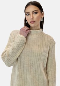 Lichtbeige geribbelde trui met een hoge hals en lange mouwen. Het materiaal lijkt zacht met een verticaal lopend, gestructureerd patroon.