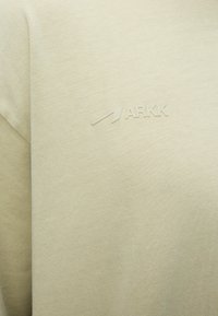 Μπεζ ύφασμα με μικρό ανάγλυφο λογότυπο που απεικονίζει ένα βέλος και το κείμενο ARKK στην περιοχή του στήθους ενός ενδύματος.