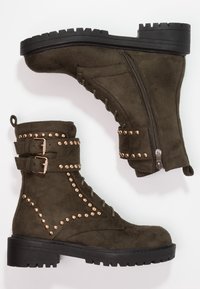 Bottines en suède vert olive avec des clous dorés, des accents de doubles boucles, une fermeture éclair sur le côté et une semelle en caoutchouc noir.