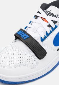 Zapatilla blanca con correa negra y acentos azules. Presenta cuero perforado, cordones planos y suela de goma. Logotipo de Nike prominentemente exhibido.
