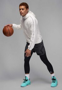 Sudadera con capucha blanca con detalles negros, pantalones cortos grises, mallas negras y zapatillas acuáticas. Sosteniendo un balón de baloncesto, en una pose dinámica.