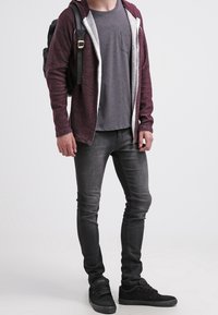 Vinröd huvtröja med vitfoder, grå t-shirt med ficka, mörka jeans och svarta sneakers. Svart ryggsäck på axeln.