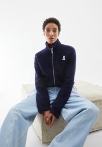 Marineblaue Fleecejacke mit hohem Kragen, Reißverschluss und Seitentaschen. Mit einem weißen Band-Logo auf der linken Brust. Kombiniert mit hellblauen, weiten Jeans.