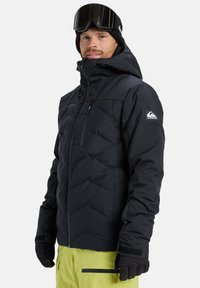Man in een zwarte winterjas met capuchon, zwarte handschoenen, een zwarte beanie, skibril en heldergele broek tegen een witte achtergrond.
