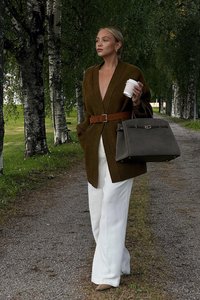 Cárdigan verde oversized con un escote en V profundo, cinturón marrón, pantalones anchos blancos y un bolso gris. Sostenido una taza de café.