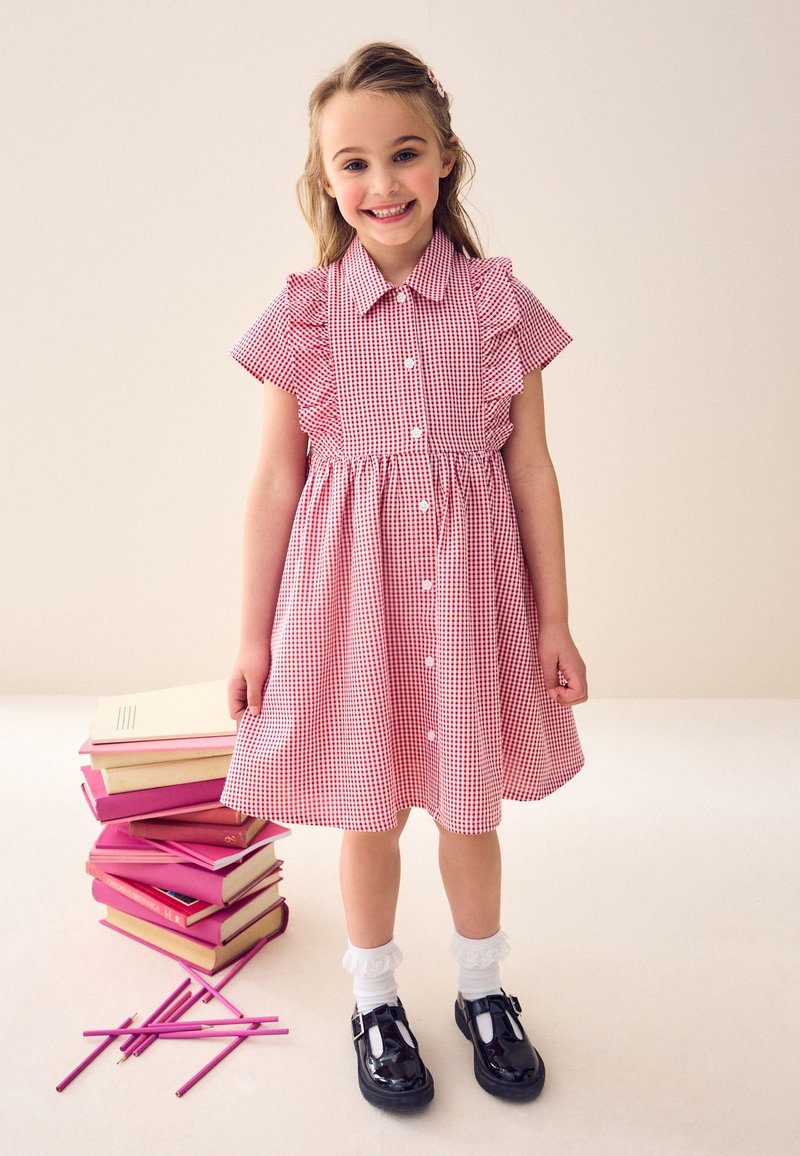 Fille souriante en robe à carreaux rouge et blanc, chaussettes blanches et chaussures noires, debout près d'une pile de livres et de crayons roses éparpillés sur le sol.