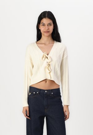 Kremfarget strikket kort cardigan med V-hals og frontløsning, med teksturerte mønstre og lange ermer. Kombinert med mørk denim jeans.