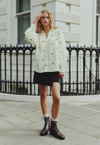Light colored, textured shirt with embroidered details, paired with a brown suede mini skirt and lace-up brown boots with white socks.