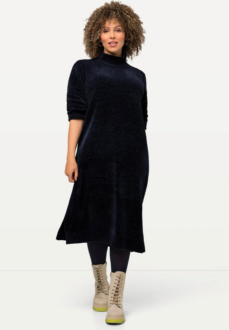 Robe-sweater longue bleu marine, tissu texturé, col haut, poignets côtelés, fentes sur les côtés ; associée à des bottes beiges à lacets et à des leggings noirs.