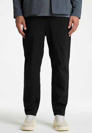 Homme portant un pantalon noir à jambes droites, une veste grise et des baskets blanches, debout les mains détendues le long du corps sur un fond blanc.