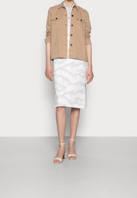 Beige utilityjack met twee borstzakken over een witte rok met een subtiel abstract patroon en een zijsplit. Transparante hakken met enkelbandje.