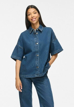 Jeune femme aux longs cheveux foncés portant une chemise en jean à manches courtes boutonnée et un pantalon en jean assorti, souriante avec une main dans la poche.