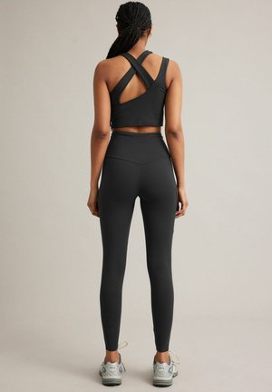 Frau mit geflochtenem Haar, die einen schwarzen Sport-BH mit Kreuzriemen, hoch taillierte Leggings und graue Sportschuhe trägt, steht mit dem Rücken zur Kamera vor einem neutralen Hintergrund.