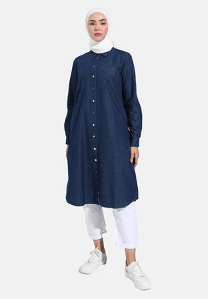 Mujer con hiyab blanco, vestido camisa largo azul oscuro abotonado, pantalones cortos blancos y zapatillas blancas, de pie contra un fondo neutro.