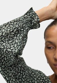 Black top with puffed sleeves, featuring a floral pattern in light green. Textured fabric with gathered detailing at the shoulder.