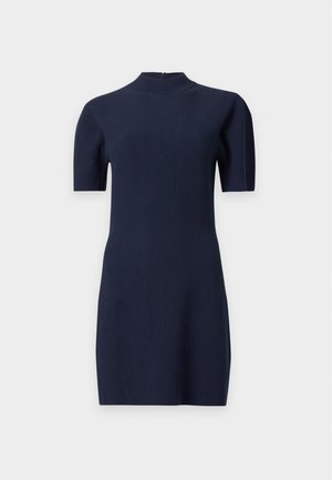 Femme debout portant une robe en maille côtelée bleu marine à manches courtes, associée à des sandales à talons noires à bout ouvert, sur un fond clair uni.