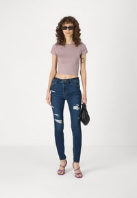 Lavas ziedu topiņš, plīstīti zilie šaurie džinsi, purpura siksnu sandales un melna rokassoma. Viļņaini mati un saulesbrilles papildina izskatu.