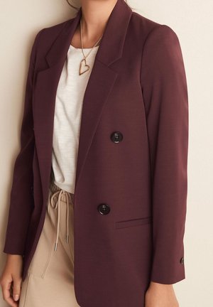Femme portant un blazer bordeaux croisé, un top blanc, un pantalon beige avec cordon et un collier avec pendentif en forme de cœur doré.