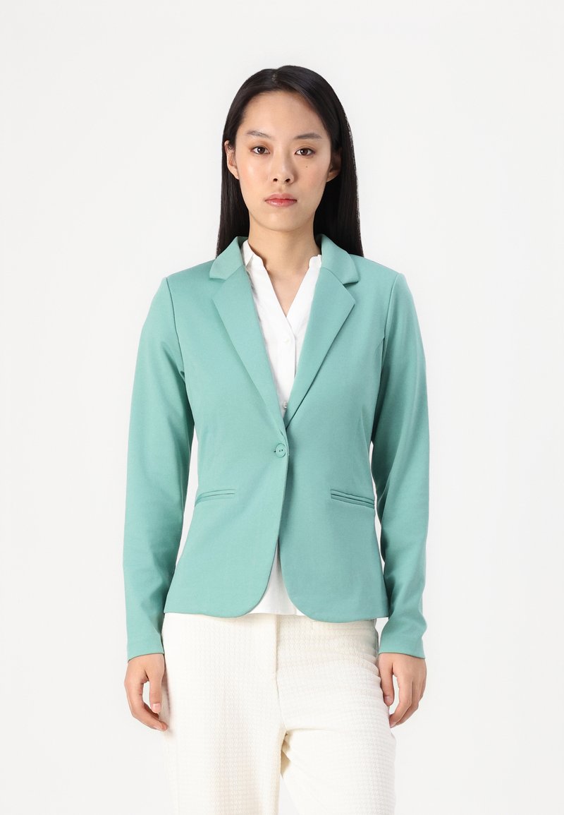 Mintgrön blazer i slätt tyg, med en knappstängning, nedskurna slag och två framfickor, tillsammans med en vit skjorta.