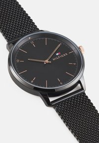 Black analog watch with a black mesh band. The face features gold-tone hour markers, hands, and "HILFIGER" logo, with a small tricolor accent.