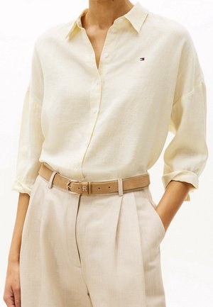 Femme portant une chemise en lin crème boutonnée avec un petit logo sur la poitrine, pantalon plissé beige taille haute et ceinture marron clair avec boucle dorée.