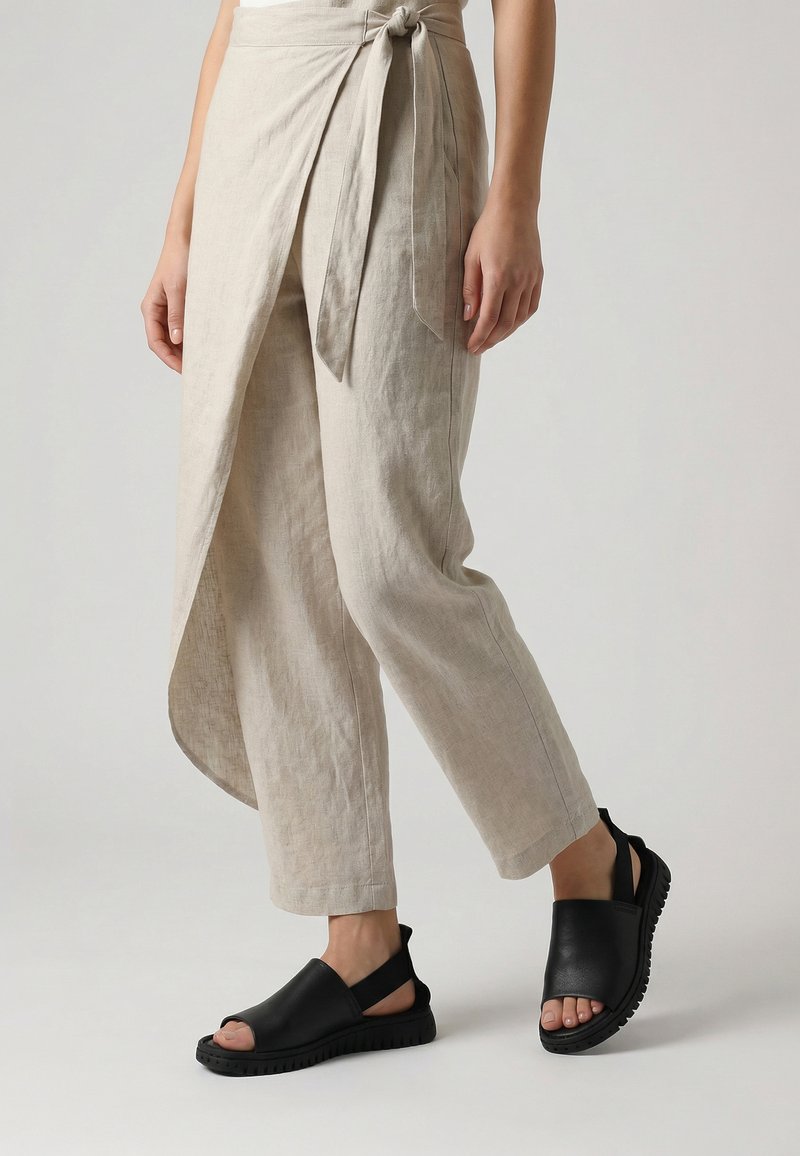 Beige bukser i omslagsstil med bindebånd i siden, båret med sorte slingback-sandaler med åbne tæer på en lys baggrund.