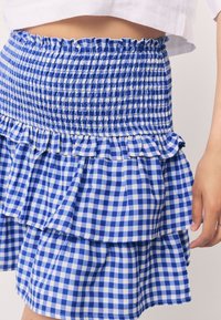 Gonna a vita alta a quadretti blu e bianchi con vita elasticizzata smock e orlo a balze a strati, indossata con un top bianco corto.