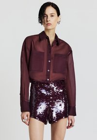 Blouse bordeaux transparent avec deux poches avant et fermeture à boutons, associée à un short ajusté recouvert de sequins violets brillants.