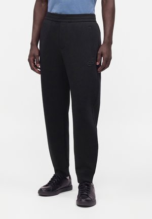 Mand iført sorte joggingbukser med elastisk talje og sidelommer, kombineret med sorte sneakers, stående mod en ensfarvet baggrund.