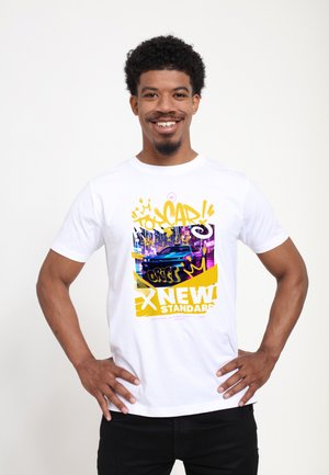 Uśmiechnięty mężczyzna z rękami na biodrach, ubrany w białą koszulkę z kolorową grafiką niebieskiego samochodu sportowego i napisem graffiti „NEW STANDARD”.