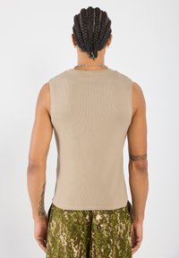 Beige ermeløs topp i ribbestrikket stoff, rund halslinning, med rett kant. Modellen har på seg mønstrede grønne camouflagebukser.