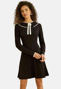 Fekete hosszú ujjú ruha pliszírozott szoknyával, hullámos szegélyű gallérral és masni díszítéssel, fehér szegéllyel az ujjakon.