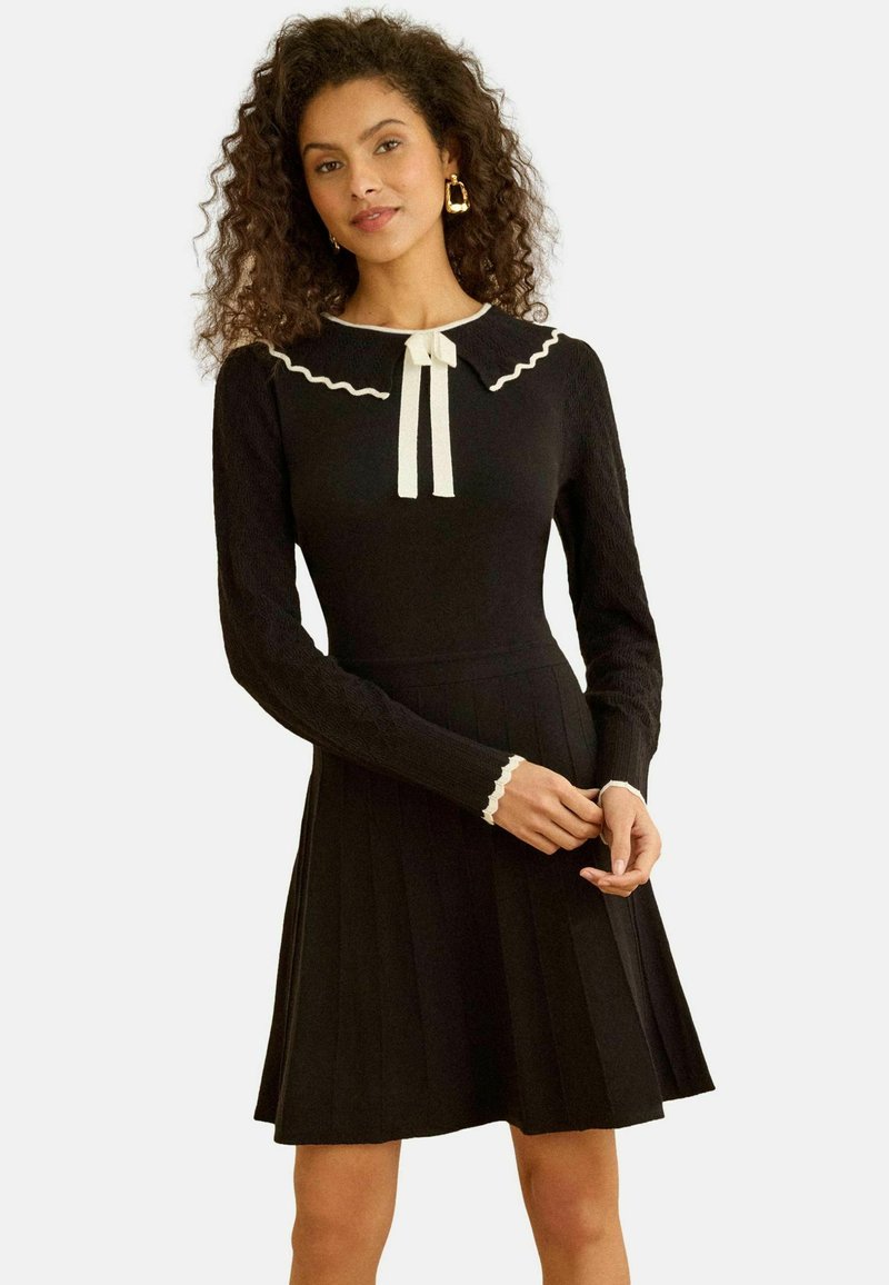 Fekete hosszú ujjú ruha pliszírozott szoknyával, hullámos szegélyű gallérral és masni díszítéssel, fehér szegéllyel az ujjakon.