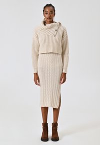 Set in maglia composto da due pezzi, con un maglione cropped crema con colletto a cascata e dettagli con bottoni, abbinato a una gonna a coste lunga fino al ginocchio.
