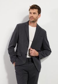 Donkergrijze blazer met notch-revers, twee knopen en zijzakken. G gedragen over een wit T-shirt, met een gladde stof en een getailleerde pasvorm.