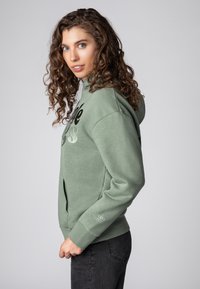 Lichtgroene hoodie met een voorkantzak, ribbelboorden en een centraal grafisch ontwerp in zwart en zilver. Zacht, textuurstof.