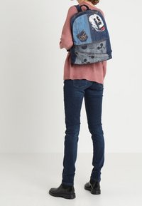 Džínový batoh s různými nášivkami, včetně grafiky postavy a kruhového vzoru. Modré tóny s texturovaným povrchem.