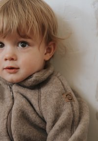 Braune Fleecejacke mit hohem Kragen und Reißverschluss. Mit einem Logo-Patch am Ärmel und sichtbaren Nahtdetails.