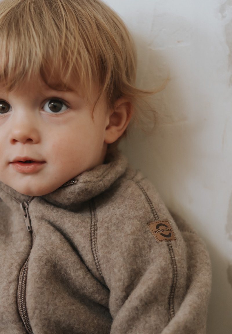 Braune Fleecejacke mit hohem Kragen und Reißverschluss. Mit einem Logo-Patch am Ärmel und sichtbaren Nahtdetails.