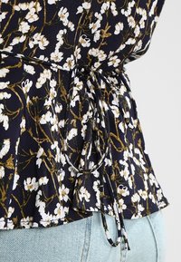 Blusa floreale blu navy con vita aderente e dettaglio a peplo. Presenta fiori bianchi e viti marroni; laccetto regolabile sul lato.