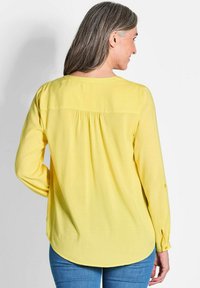 Camicia gialla a maniche lunghe con scollatura rotonda, dettaglio plissettato sul retro, polsini con bottone e vestibilità morbida, abbinata a jeans in denim.