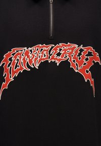 Črno oblačilo s srebrnim polovičnim zadrgo in rdečim robom z nazobčanimi črkami "SAVAGE" izvezeno čez prsni del.