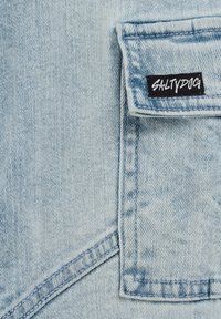 Svetlo modra denim tkanina s žepom na zakrpi, z črnim oznako z napisom "SALT DOG" v belem besedilu. Vidne šive in podrobnosti teksture.